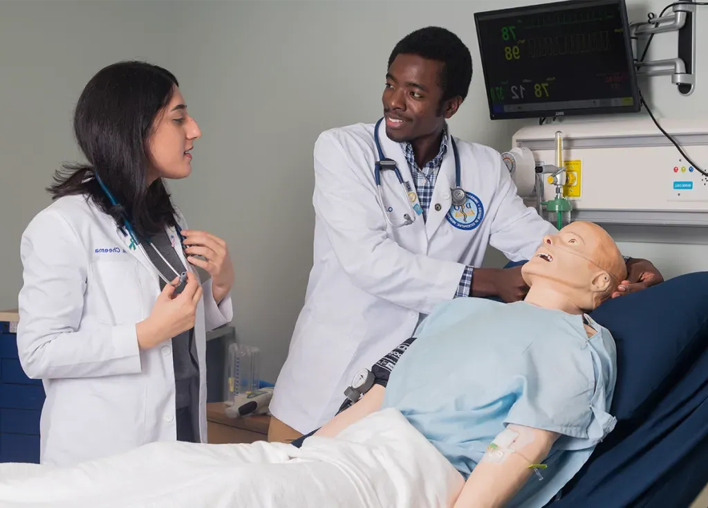 Two C O M students, in their branded white coats, work with a patient simulator